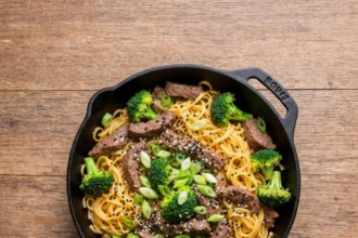 Serving Stage: Plated beef and broccoli noodle skillet on a white plate, garnished with sesame seeds and scallions, lime wedge on the side, bright and inviting presentation, ready to eat.